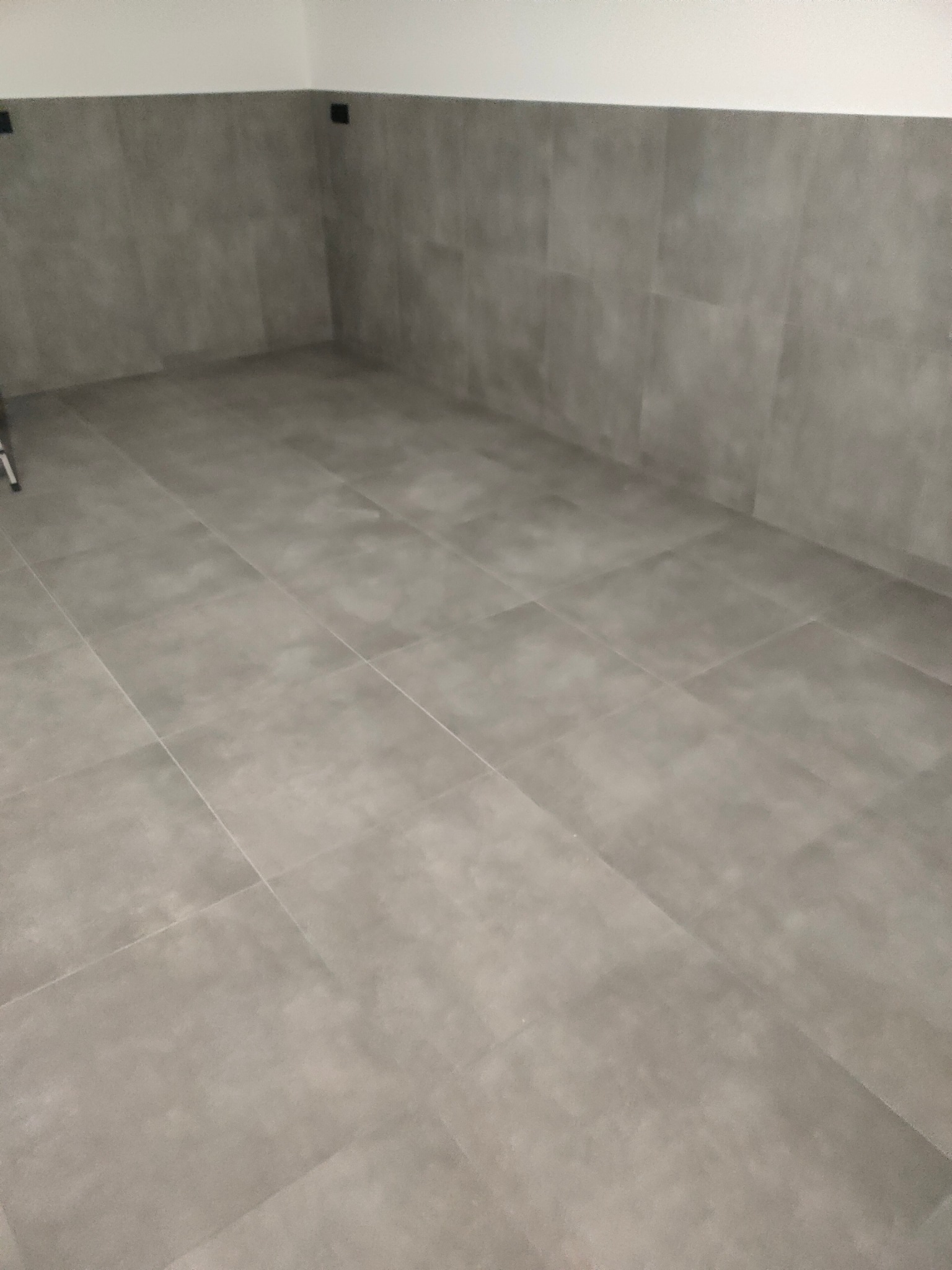 Szare, matowe płytki podłogowe i ścienne w pustym pomieszczeniu. Minimalistyczny design, widoczne fugi i gniazdka elektryczne na ścianie. Ukończony remont łazienki.