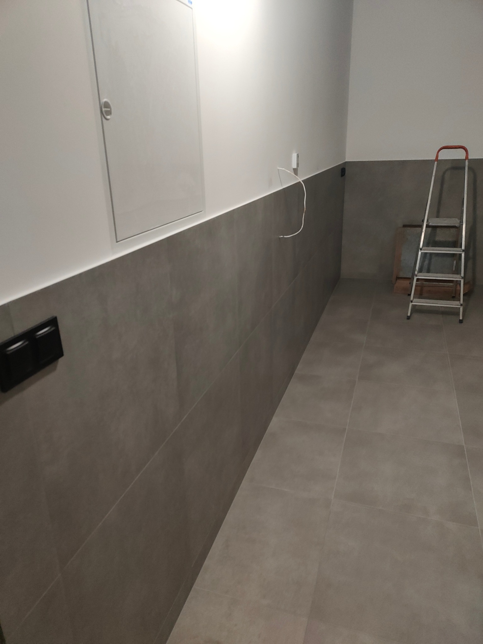 Wykończona łazienka z szarą glazurą do połowy ściany, białe ściany powyżej. Widoczna drabina malarska, gniazdka i wystające kable. Minimalistyczny design.