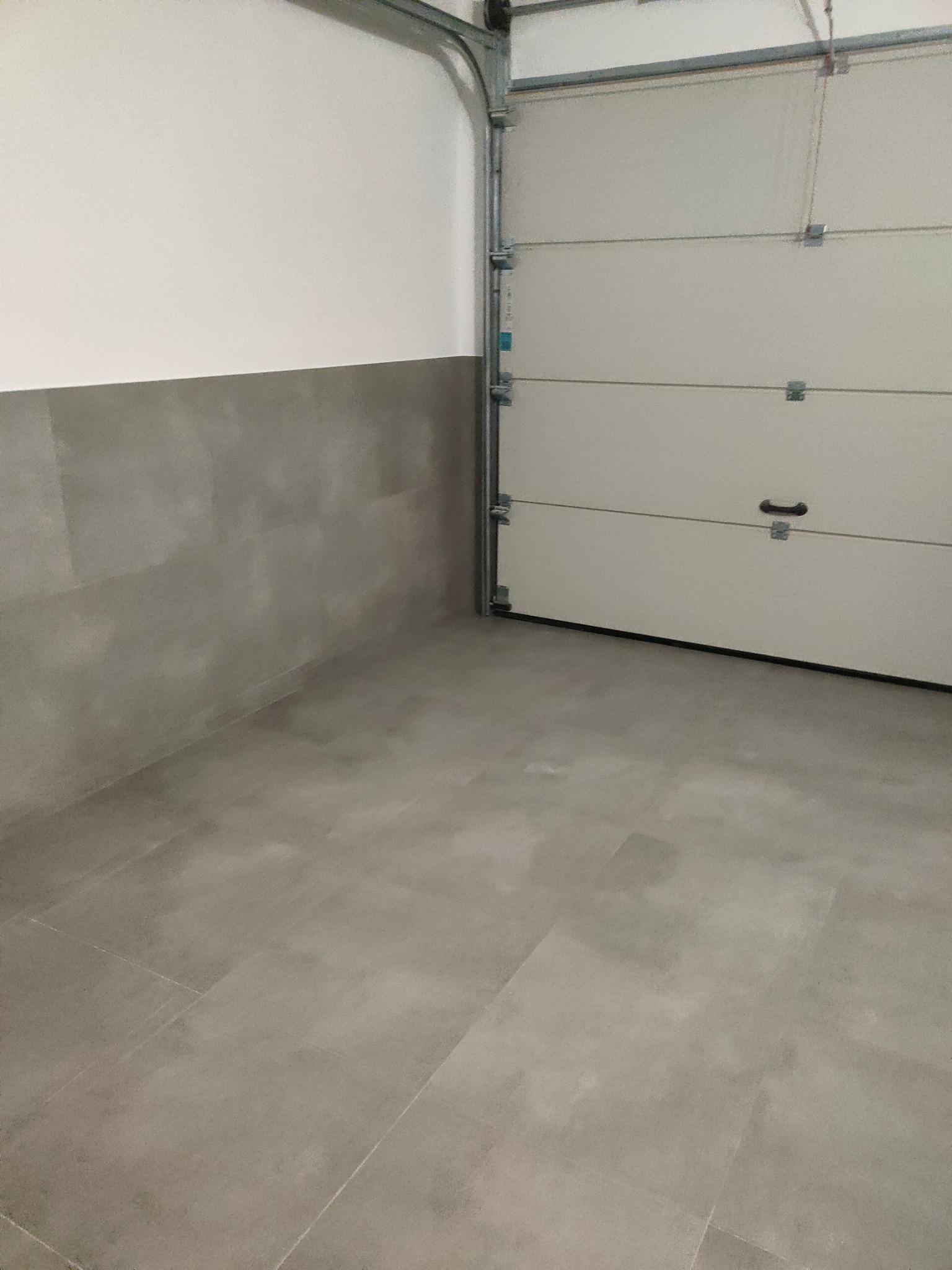 Wykończone wnętrze garażu z szarą podłogą z płytek i częściowo wykończonymi ścianami. Widoczna brama garażowa.