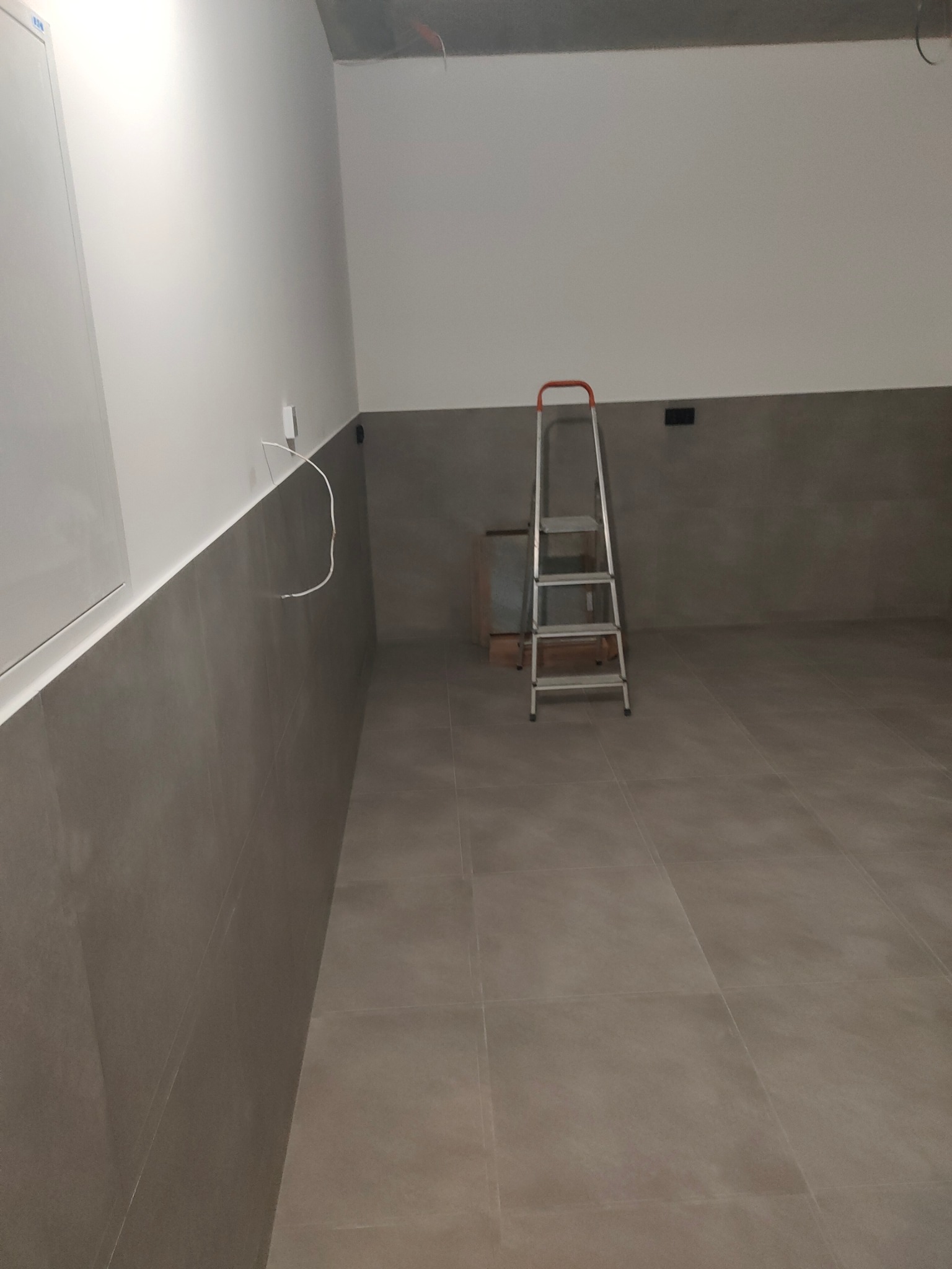 Wnętrze w trakcie wykańczania: szare płytki podłogowe i ścienne, białe ściany, drabina aluminiowa. Widoczne przewody elektryczne. Minimalistyczny, surowy styl.