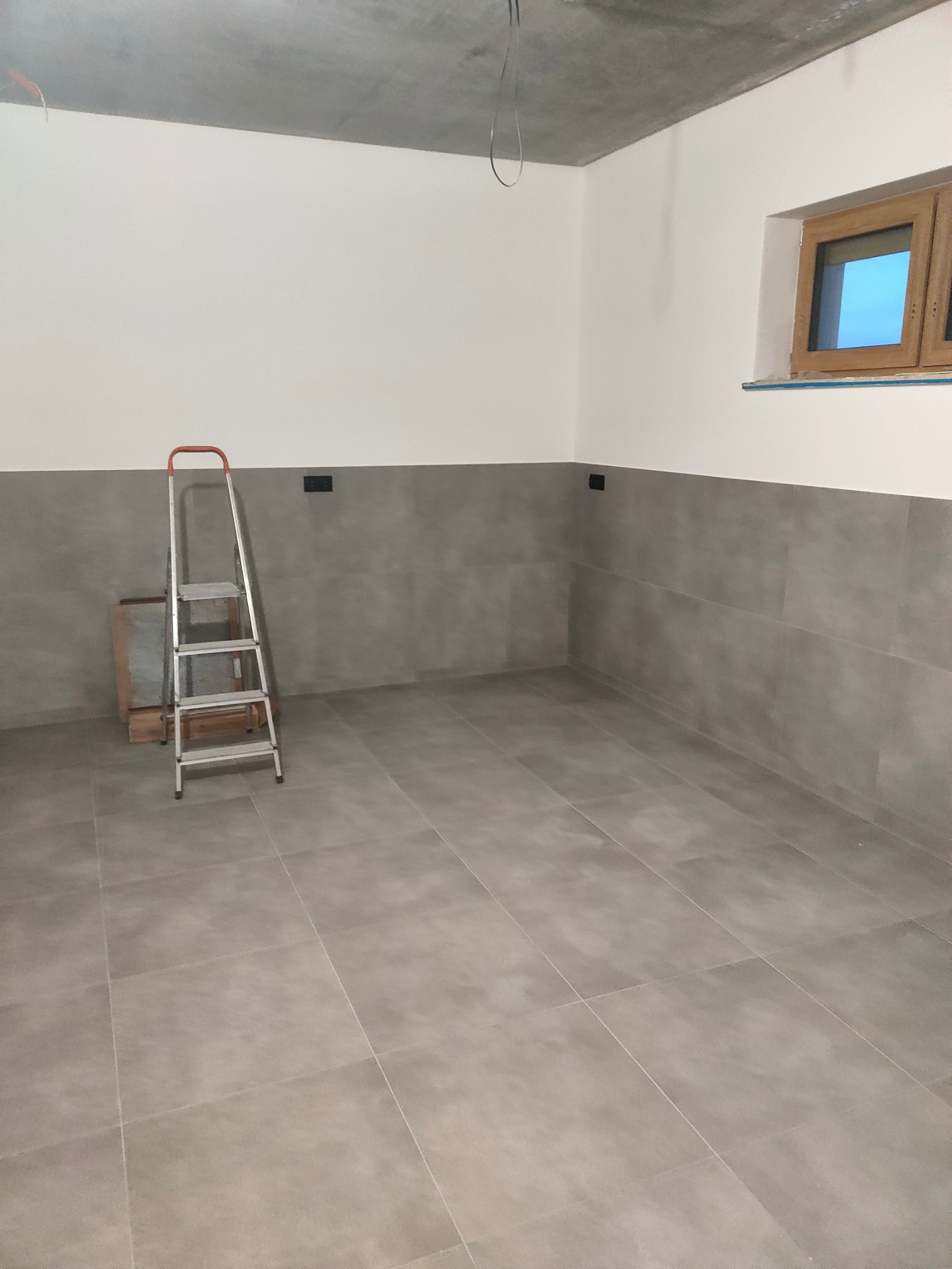 Szara, wykafelkowana łazienka w trakcie remontu. Widoczna drabina, okno i surowy sufit. Płytki na podłodze i ścianach do połowy wysokości.