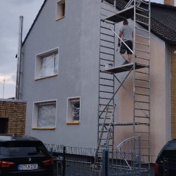 Usługi remontowo-budowlane MICHAŁ KORNACKI - Pracownik na rusztowaniu maluje elewację domu jednorodzinnego na szaro, okna zabezpieczone folią, widoczny fragment samochodu z przodu posesji.