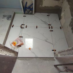 Usługi remontowo-budowlane MICHAŁ KORNACKI - Układanie białych płytek imitujących marmur na podłodze w łazience, widoczne krzyżyki dystansowe, paczka z klipsami poziomującymi i fragment instalacji hydraulicznej w suficie.