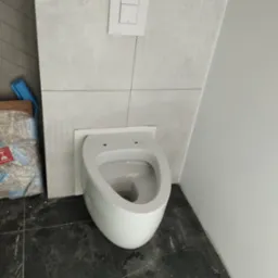 Podwieszana biała miska WC w trakcie montażu, widoczne otwory montażowe, obok opakowanie po materiałach budowlanych, ciemna podłoga z płytek.