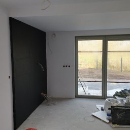 Usługi remontowo-budowlane MICHAŁ KORNACKI - Wnętrze w trakcie prac wykończeniowych: czarna ściana z izolacją, wystające kable elektryczne, surowa podłoga, okno z widokiem na zewnątrz i materiały budowlane w tle.