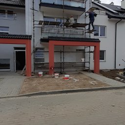 Usługi remontowo-budowlane MICHAŁ KORNACKI - Dwóch pracowników na rusztowaniu przy białym budynku z czerwonymi elementami, widoczne puszki z farbą na ziemi, prawdopodobnie w trakcie prac wykończeniowych elewacji.