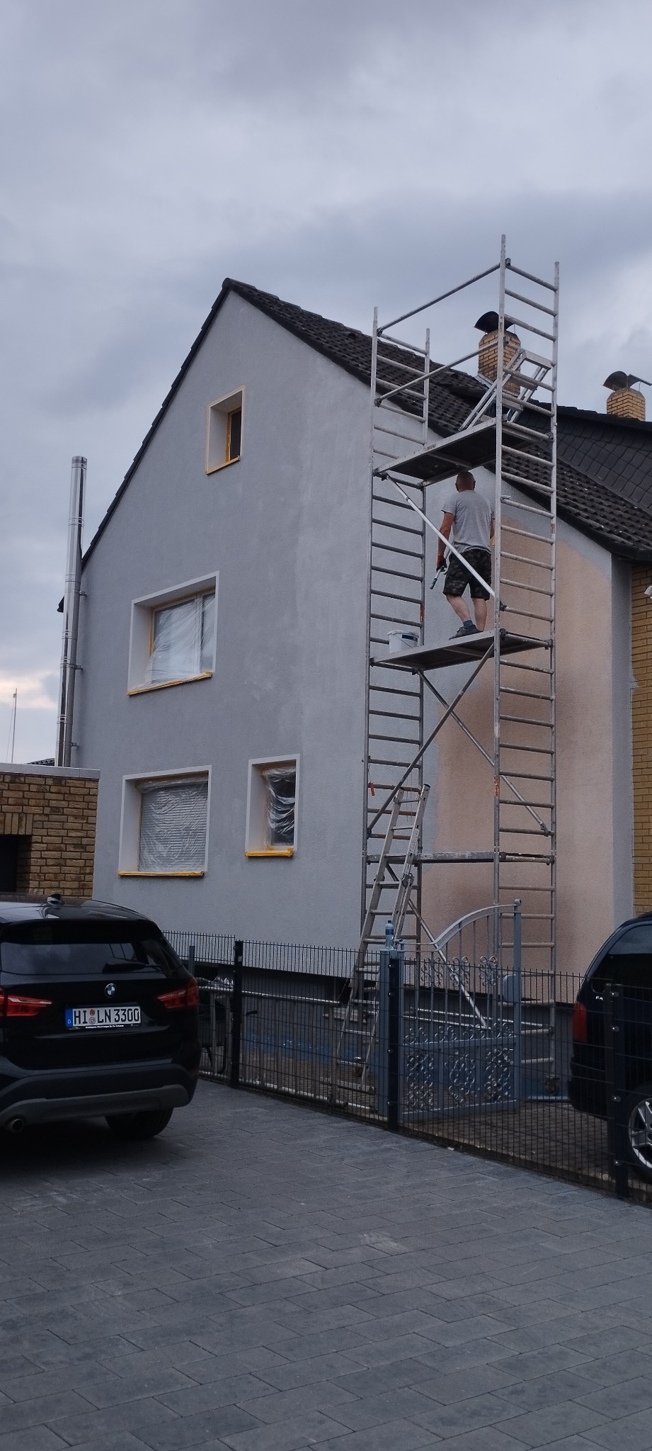Pracownik na rusztowaniu maluje elewację domu jednorodzinnego na szaro, okna zabezpieczone folią, widoczny fragment samochodu z przodu posesji.