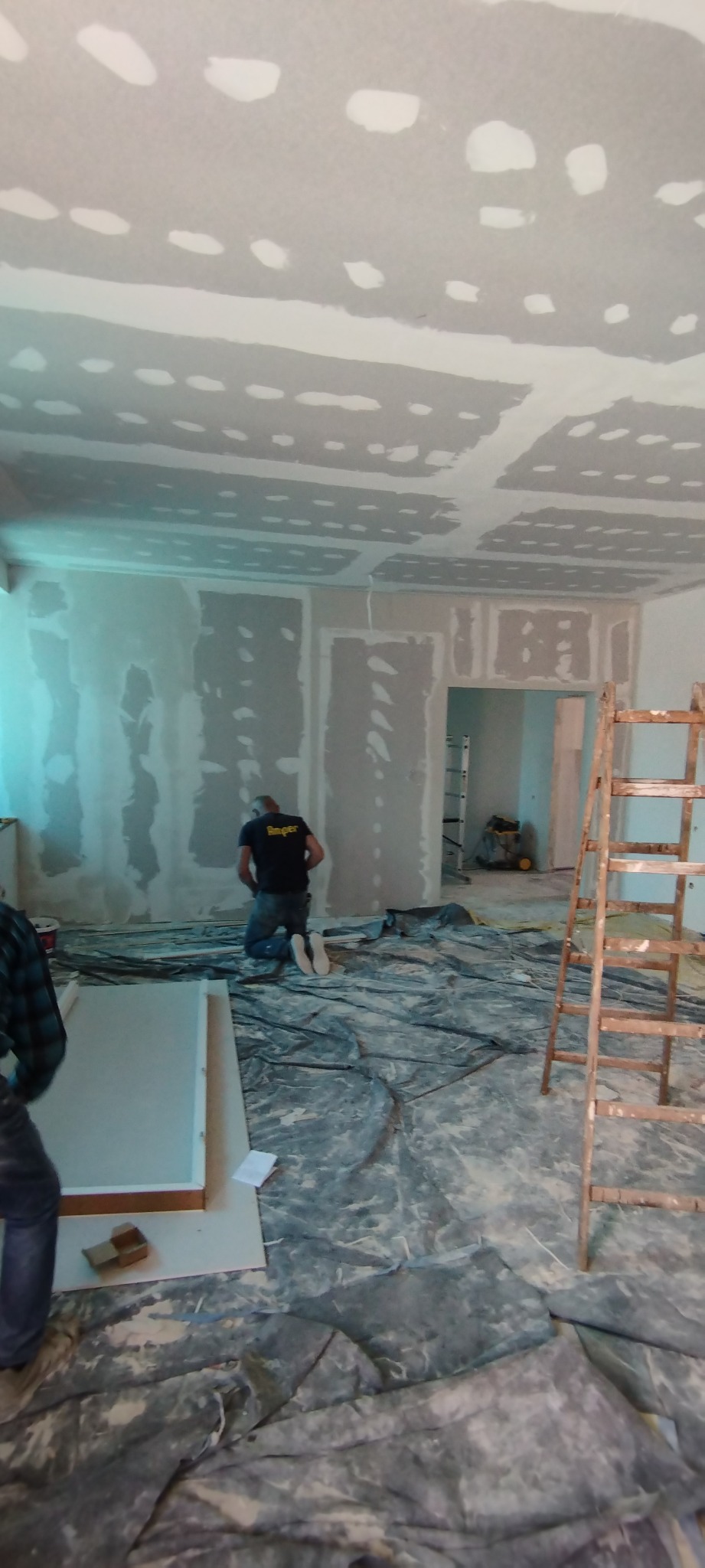 Wnętrze w trakcie prac wykończeniowych: ściany i sufit pokryte płytami gipsowo-kartonowymi, widoczne łączenia wymagające szpachlowania, pracownik w ciemnej koszulce na kolanach, rozłożona folia...