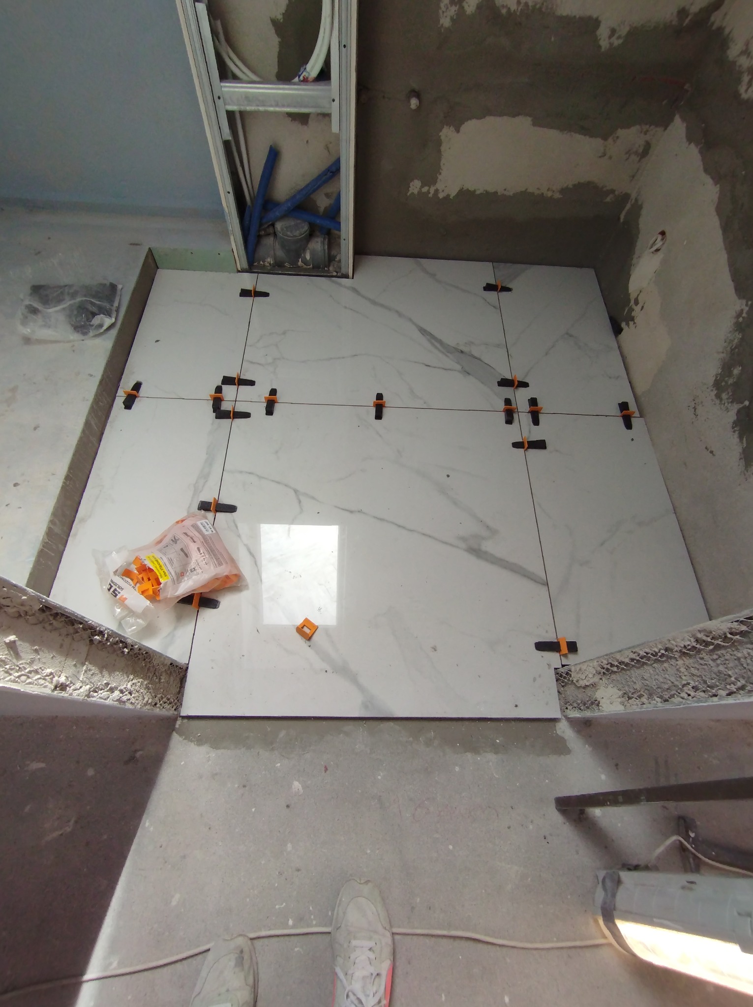 Układanie białych płytek imitujących marmur na podłodze w łazience, widoczne krzyżyki dystansowe, paczka z klipsami poziomującymi i fragment instalacji hydraulicznej w suficie.