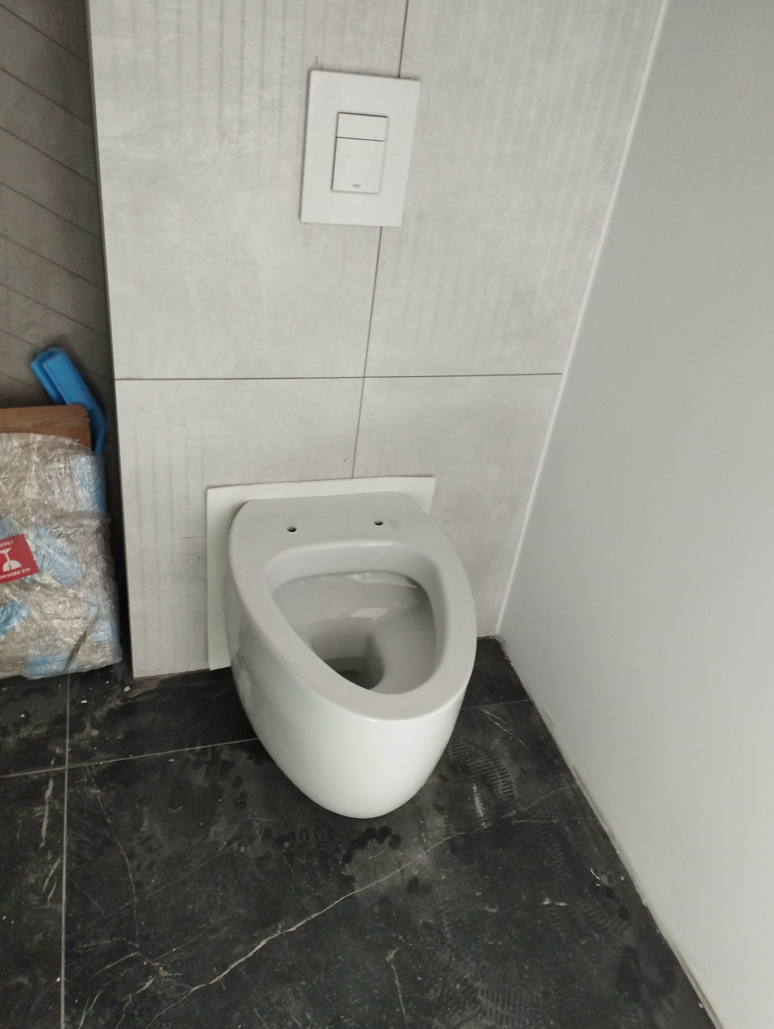 Podwieszana biała miska WC w trakcie montażu, widoczne otwory montażowe, obok opakowanie po materiałach budowlanych, ciemna podłoga z płytek.