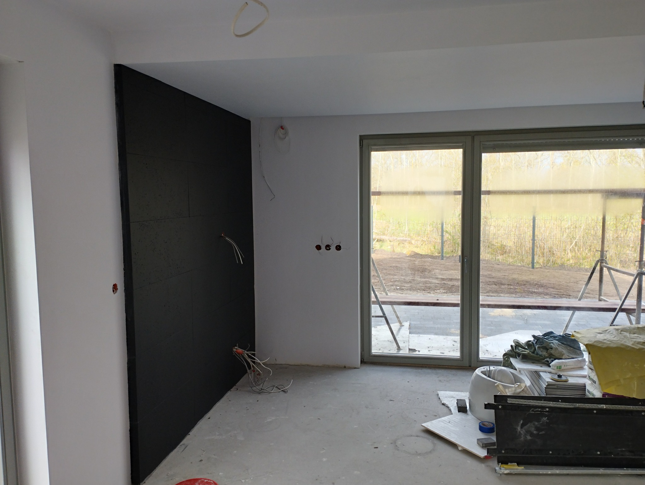 Wnętrze w trakcie prac wykończeniowych: czarna ściana z izolacją, wystające kable elektryczne, surowa podłoga, okno z widokiem na zewnątrz i materiały budowlane w tle.