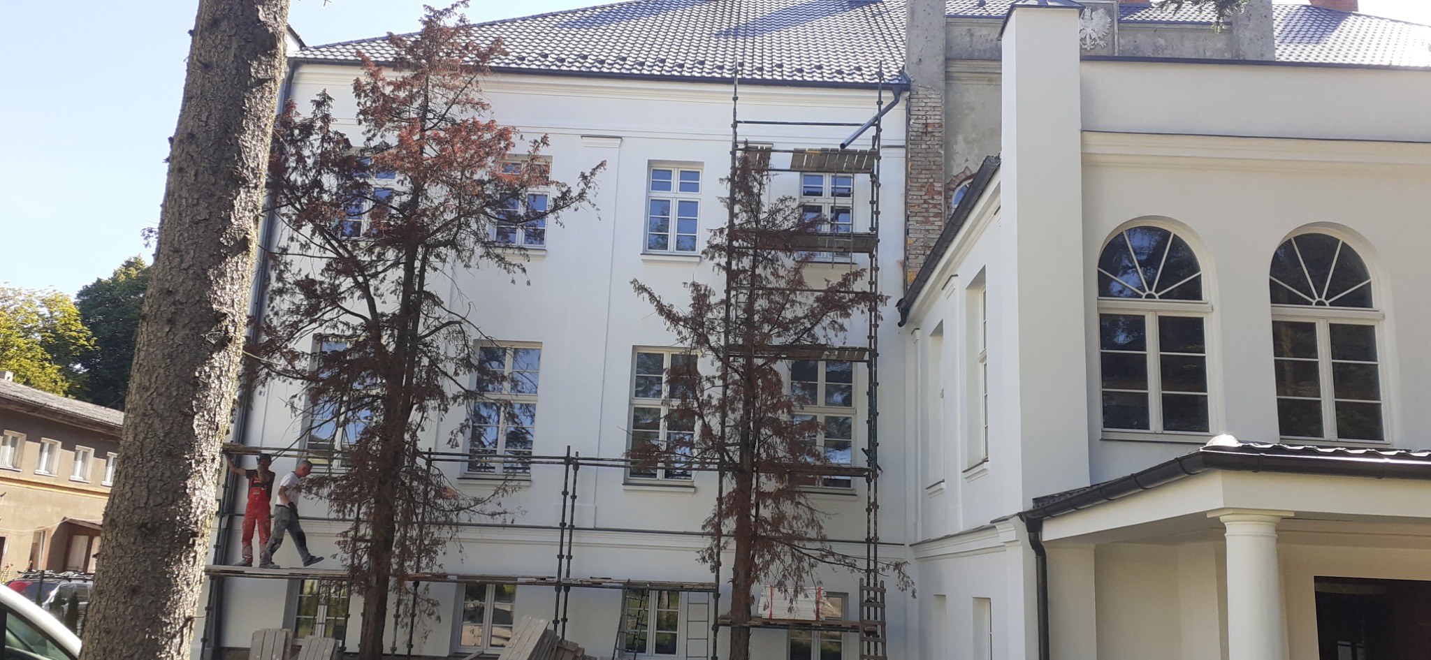 Dwóch pracowników na rusztowaniu przy białej elewacji budynku z widocznymi oknami, w tle drzewa.