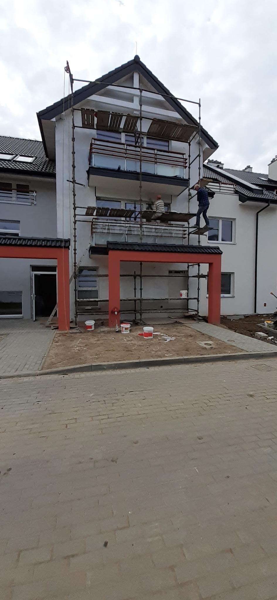 Dwóch pracowników na rusztowaniu przy białym budynku z czerwonymi elementami, widoczne puszki z farbą na ziemi, prawdopodobnie w trakcie prac wykończeniowych elewacji.