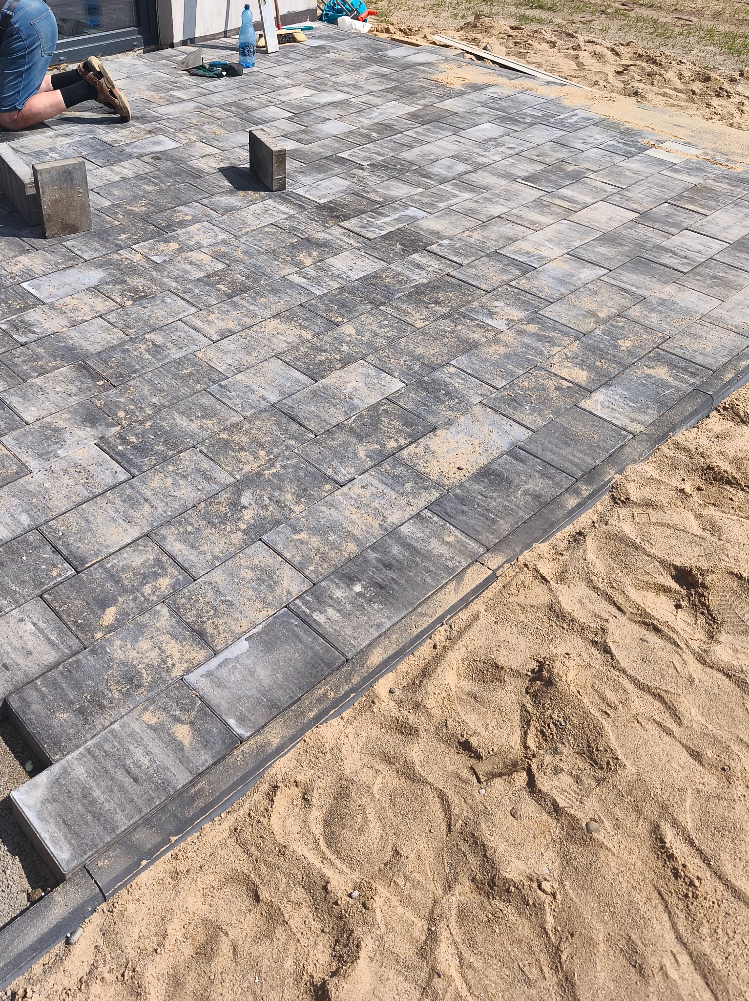 Układanie kostki brukowej: szare, prostokątne elementy z piaskiem w szczelinach, widoczne krawężniki i przygotowane podłoże z piasku. Częściowo ułożona nawierzchnia.
