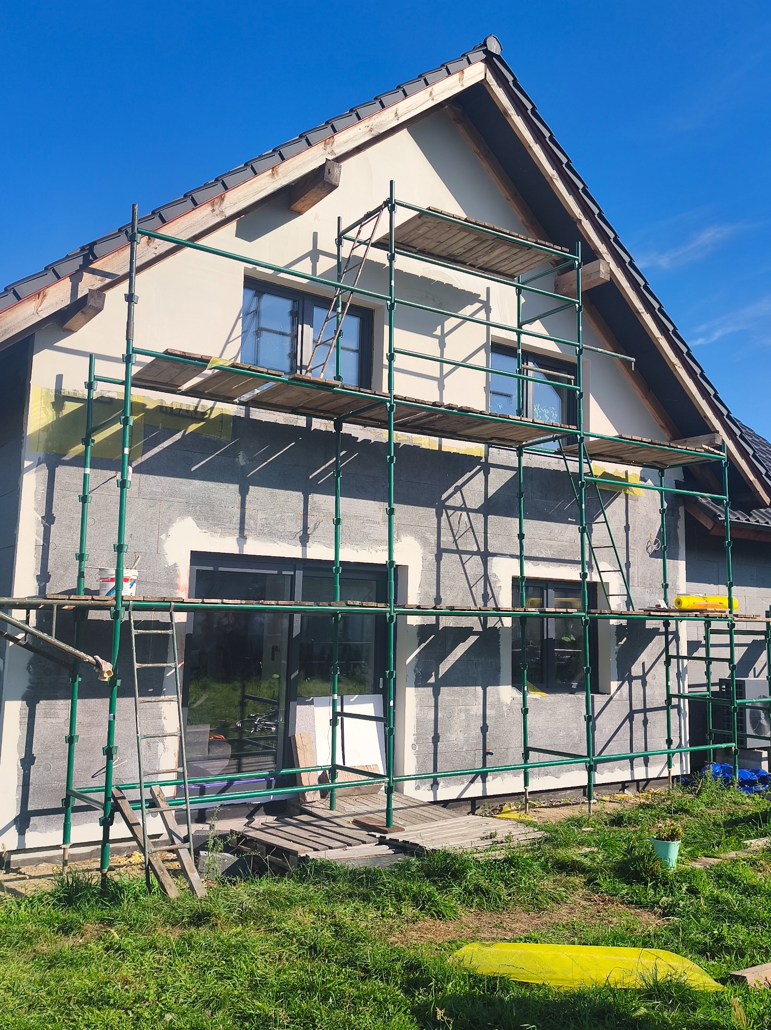 Dom jednorodzinny w trakcie ocieplania, z widocznym styropianem i rusztowaniem, na tle błękitnego nieba. Częściowo otynkowana fasada.