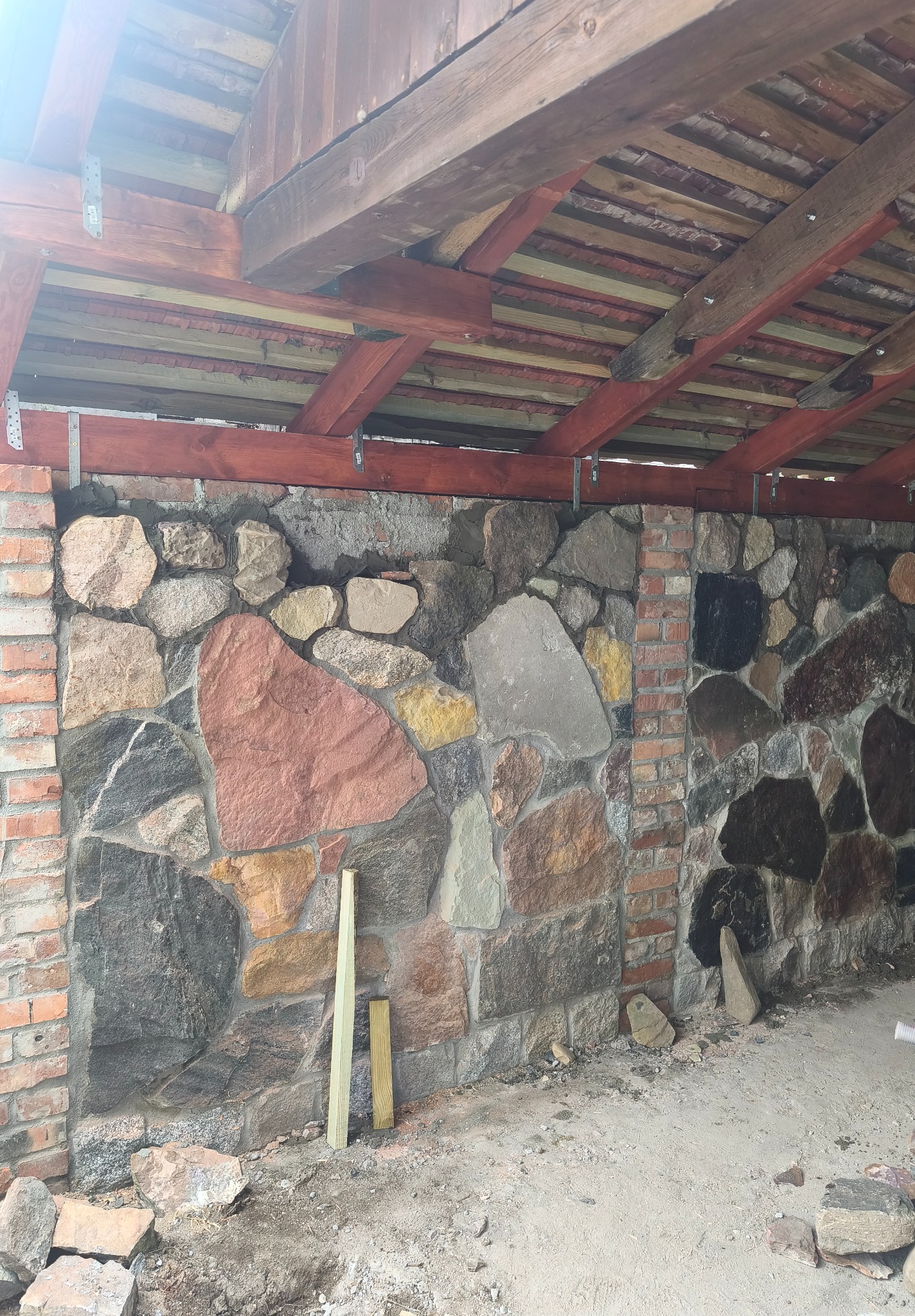 Kamienna ściana z ceglanymi kolumnami pod drewnianym zadaszeniem, widoczne fragmenty zaprawy, surowe wykończenie, dwa drewniane elementy leżące przy ścianie.