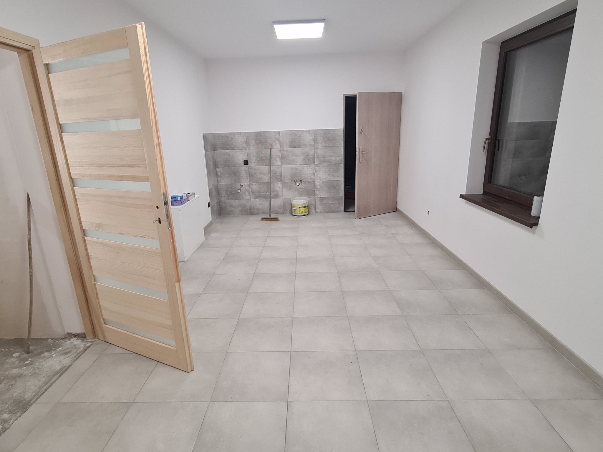 Wnętrze w trakcie remontu: szare płytki na podłodze i ścianie, otwarta drewniana ościeżnica drzwiowa z szybami, widoczny fragment pędzla, wiadro z zaprawą i mop przy ścianie z płytkami.