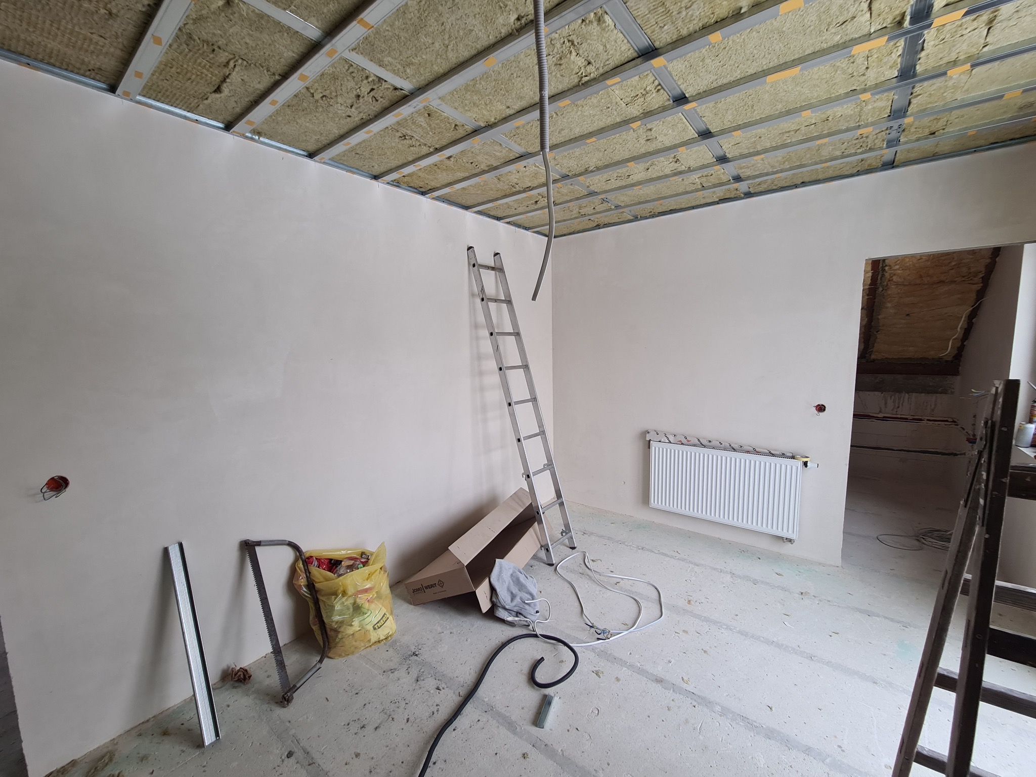 Wnętrze w trakcie prac wykończeniowych: sufit z metalową konstrukcją i izolacją z wełny mineralnej, gładkie ściany, grzejnik, drabina aluminiowa, przewody elektryczne i narzędzia na podłodze.