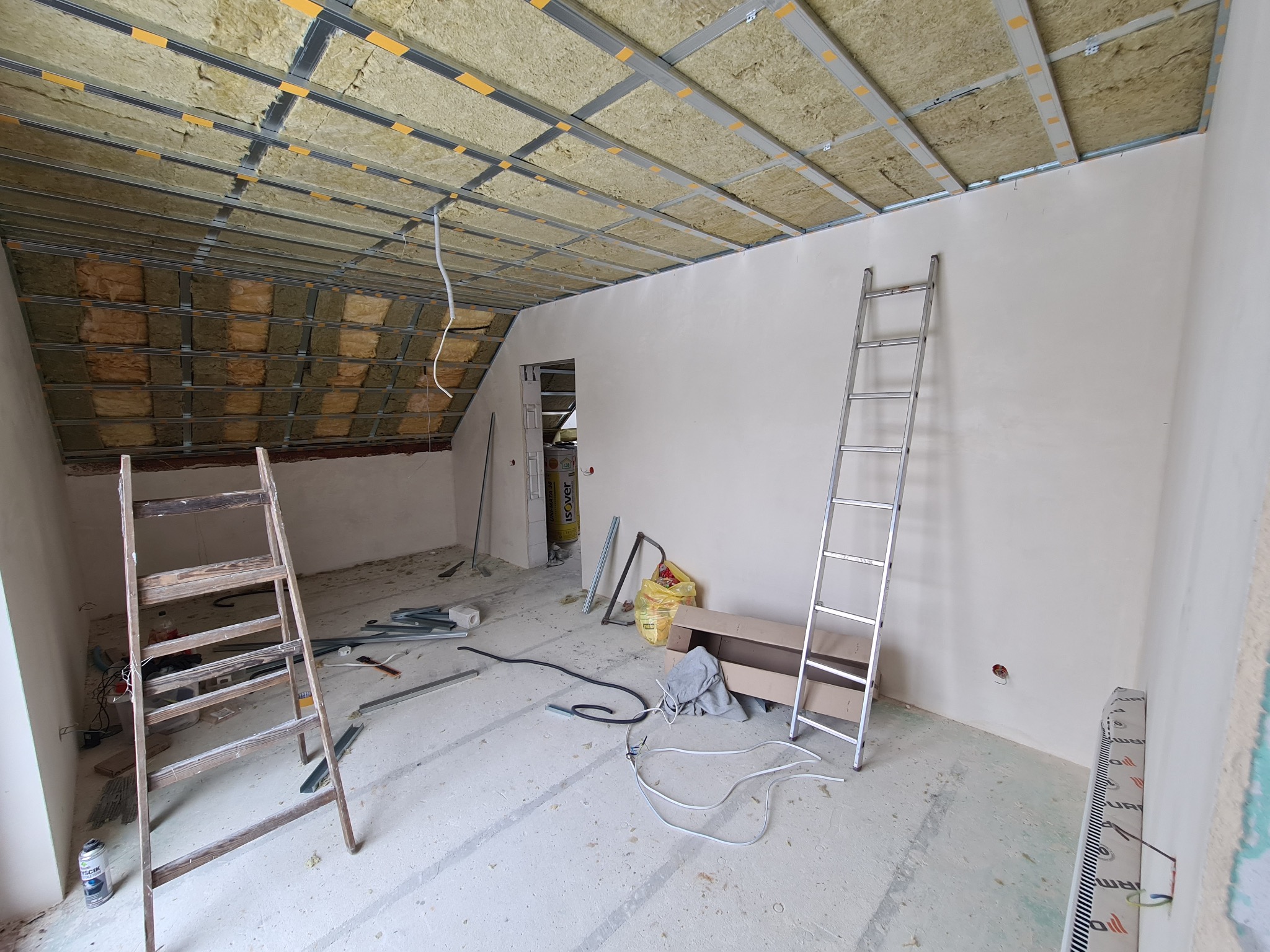 Poddasze w trakcie adaptacji: widoczne ocieplenie wełną mineralną pod metalowym stelażem sufitowym, dwie drabiny (drewniana i aluminiowa), przewody elektryczne na podłodze, resztki materiałów...