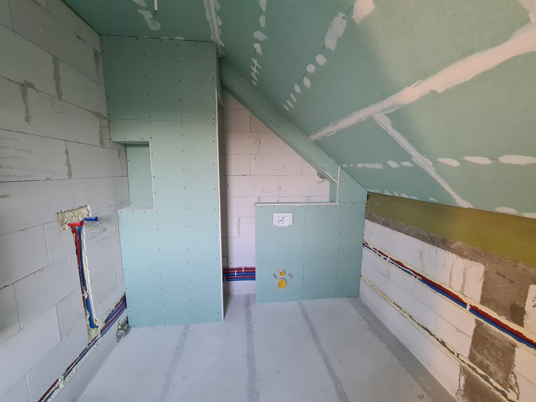 Wnętrze poddasza w trakcie adaptacji na łazienkę: ściany z bloków betonowych z instalacją wodną, zabudowa z zielonych płyt kartonowo-gipsowych, widoczne łączenia płyt wymagające szpachlowania.