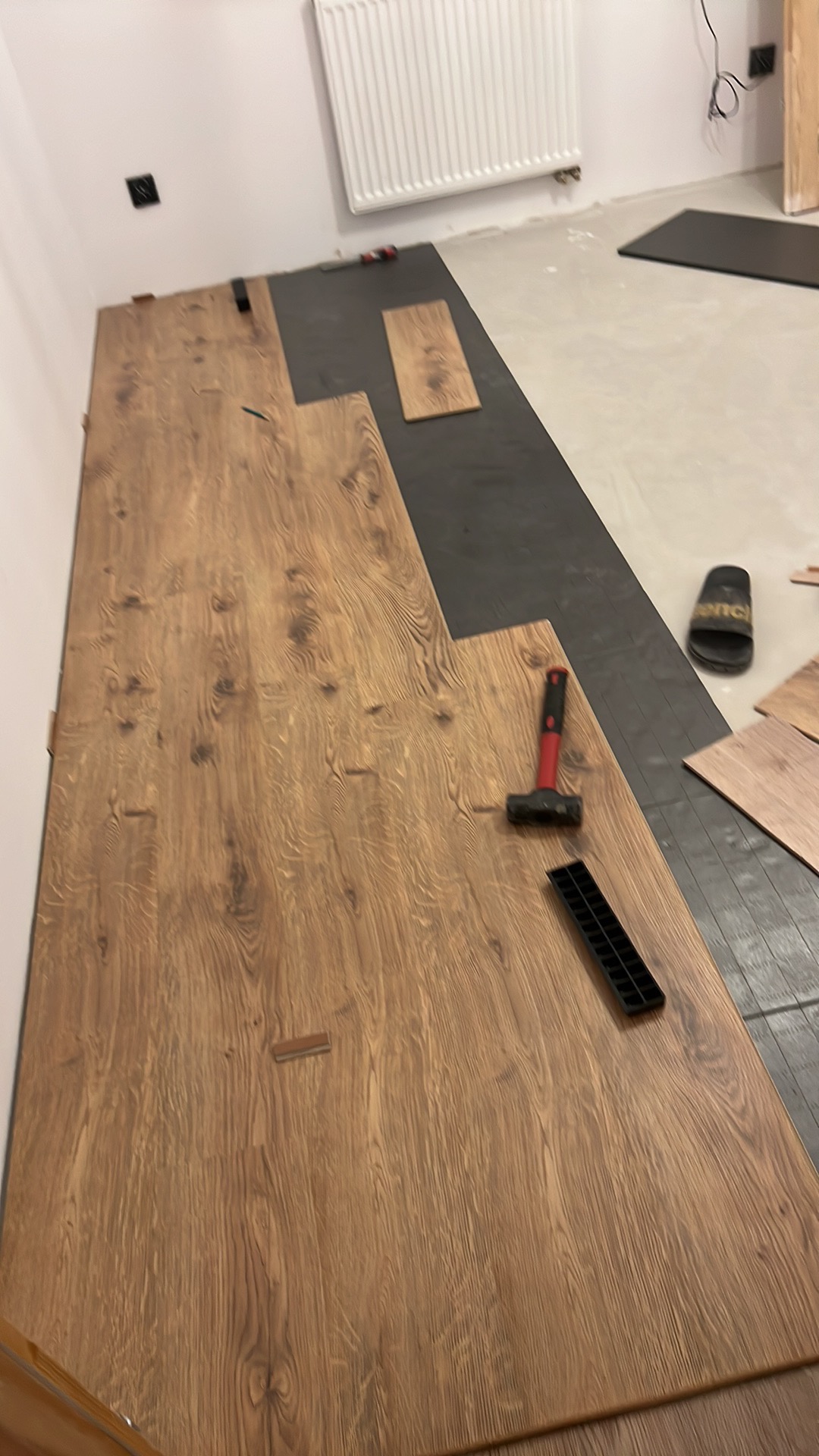 Układanie paneli podłogowych w trakcie realizacji, widoczny młotek, listwa montażowa, podkład i fragment ułożonej podłogi z widocznym wzorem drewna.