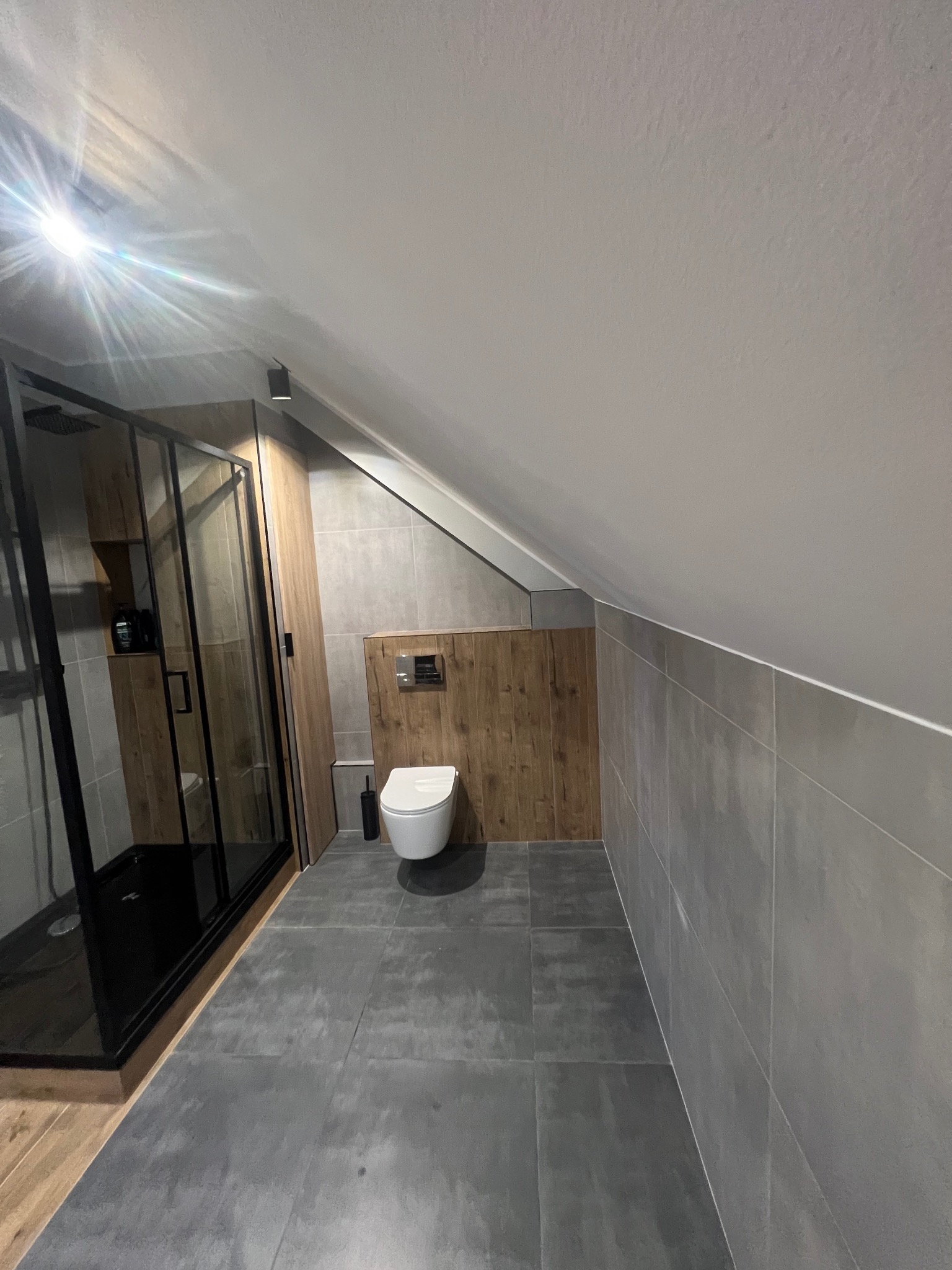 Nowoczesna łazienka na poddaszu z kabiną prysznicową typu walk-in z czarną ramą, wiszącą toaletą i płytkami imitującymi drewno na ścianie przy zabudowie stelaża podtynkowego.