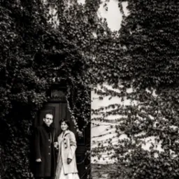 Czarno-białe zdjęcie pary narzeczeńskiej stojącej na schodach przed drzwiami obrośniętymi bluszczem, kobieta w sukni ślubnej i płaszczu, mężczyzna w ciemnym płaszczu.