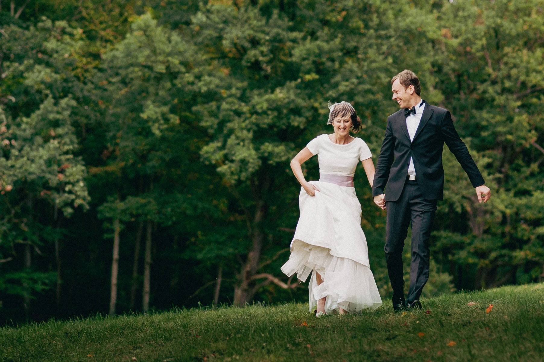 Uśmiechnięta para młoda, on w czarnym garniturze z muchą, ona w białej sukni z fioletową szarfą i woalką, trzymają się za ręce i biegną po zielonej trawie na tle drzew.