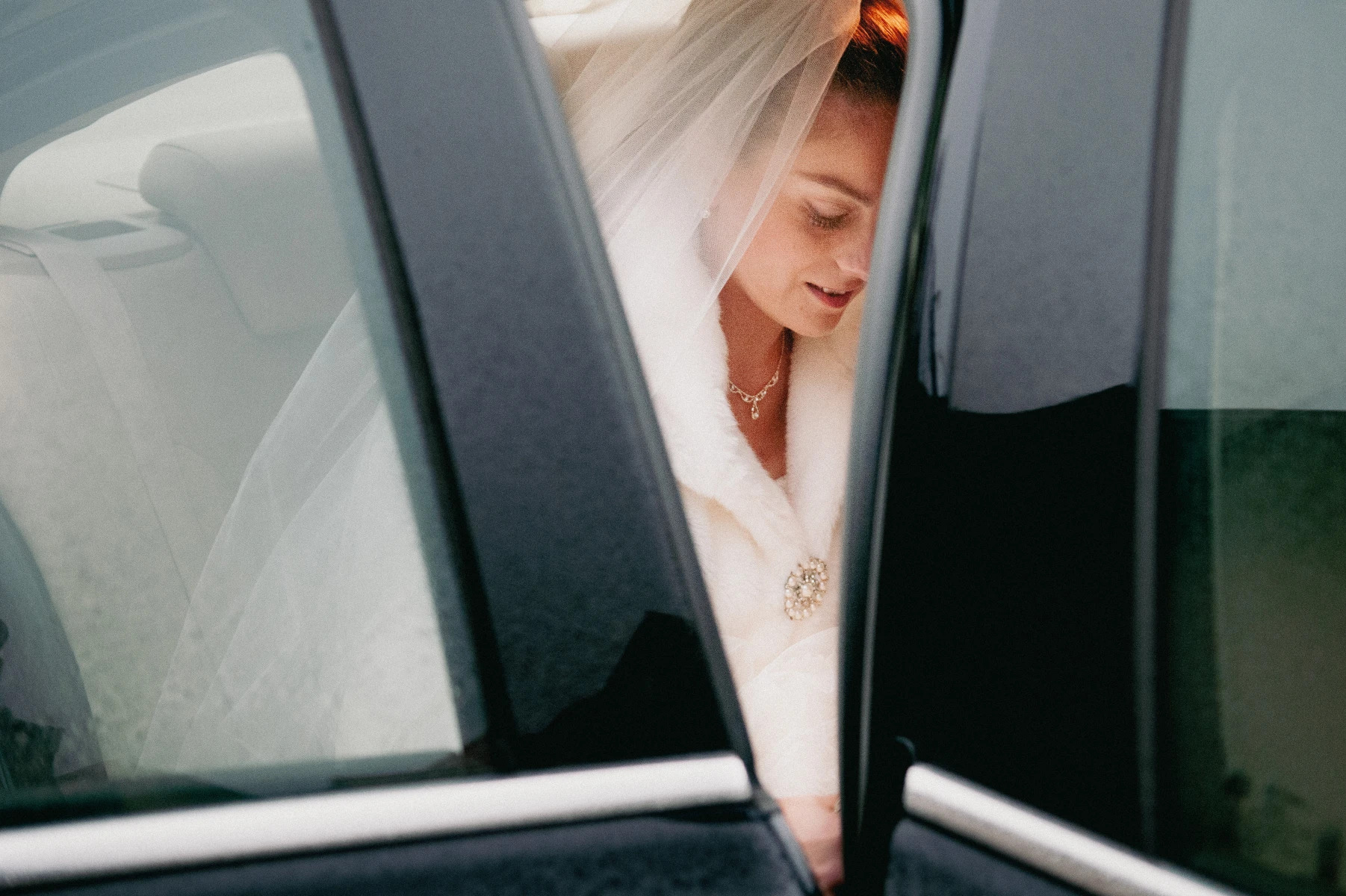 Panna młoda w welonie i futrzanym okryciu, widoczna przez otwarte drzwi czarnego samochodu, z delikatnym uśmiechem spogląda w dół.