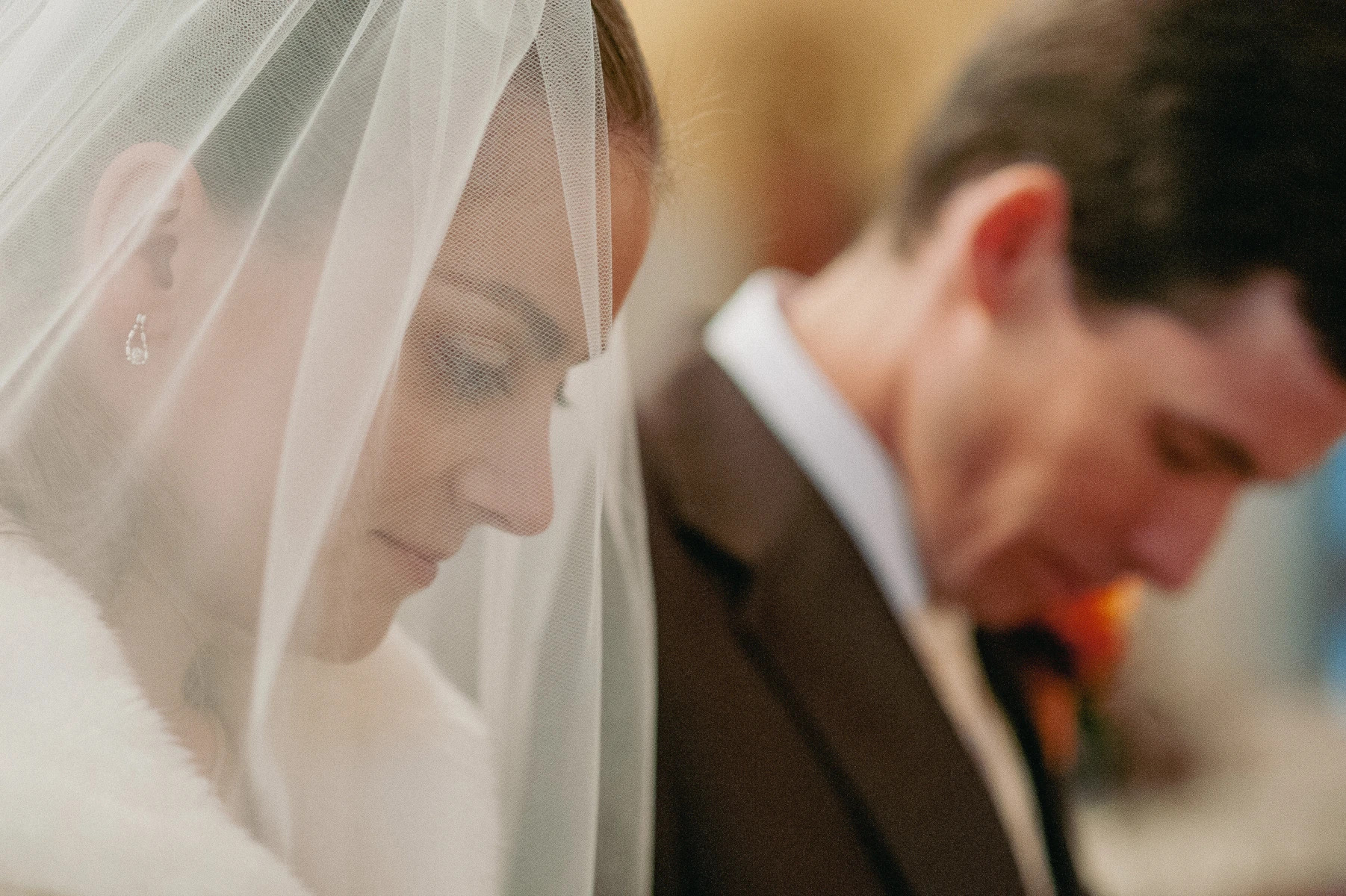 Panna młoda w białym welonie z delikatnym makijażem i łzą w oku, obok pochylony pan młody w ciemnym garniturze, intymny moment podczas ceremonii ślubnej.