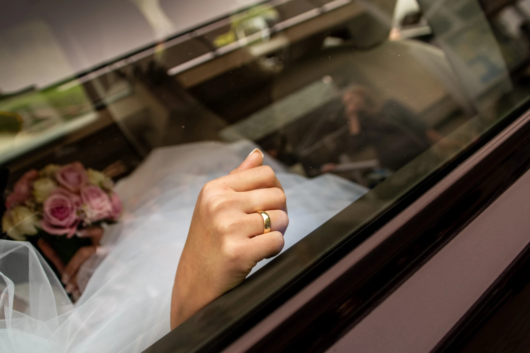Panna młoda w sukni ślubnej, z bukietem różowych kwiatów, wystawia dłoń z złotą obrączką przez okno samochodu w Poznaniu. Widoczne odbicie fotografa w szybie.