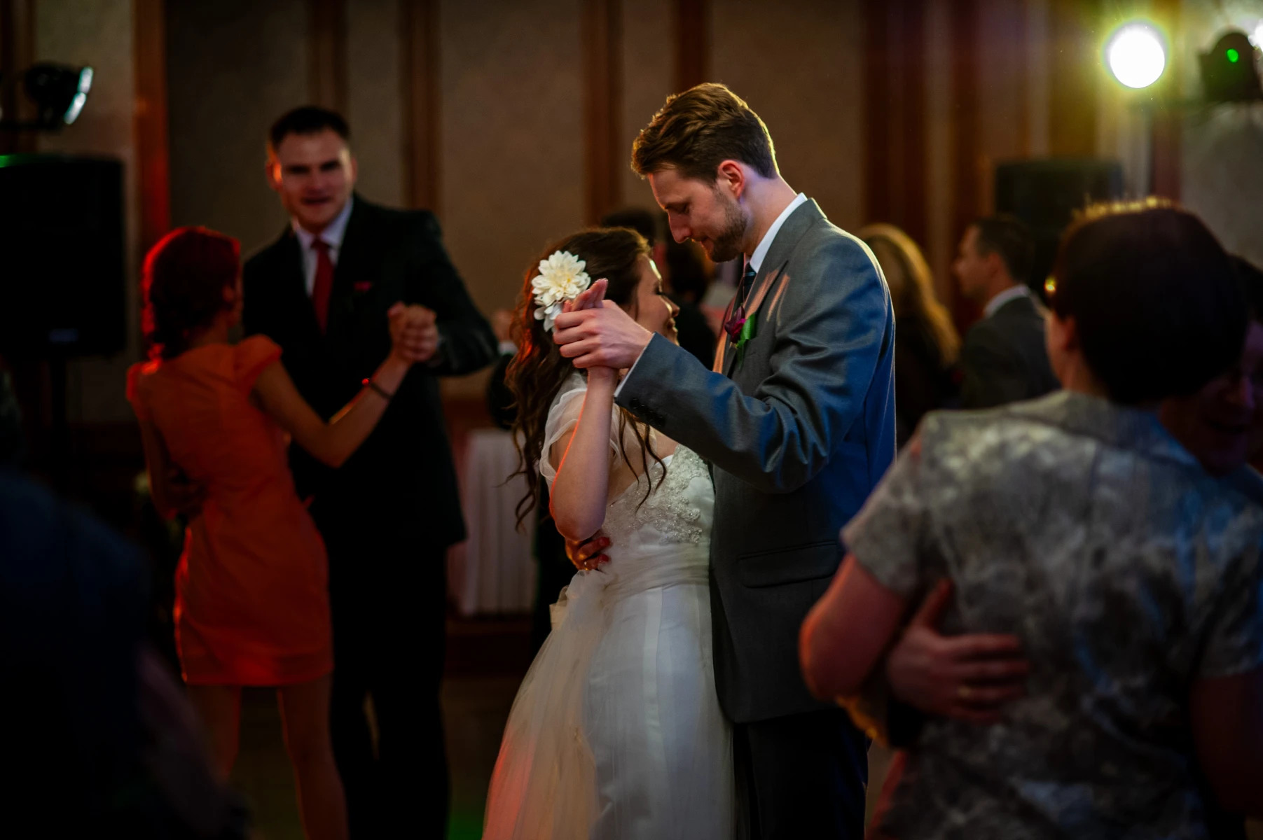 Para młoda tańczy na weselu, panna młoda w białej sukni z kwiatem we włosach, pan młody w szarym garniturze, w tle inni goście na parkiecie.