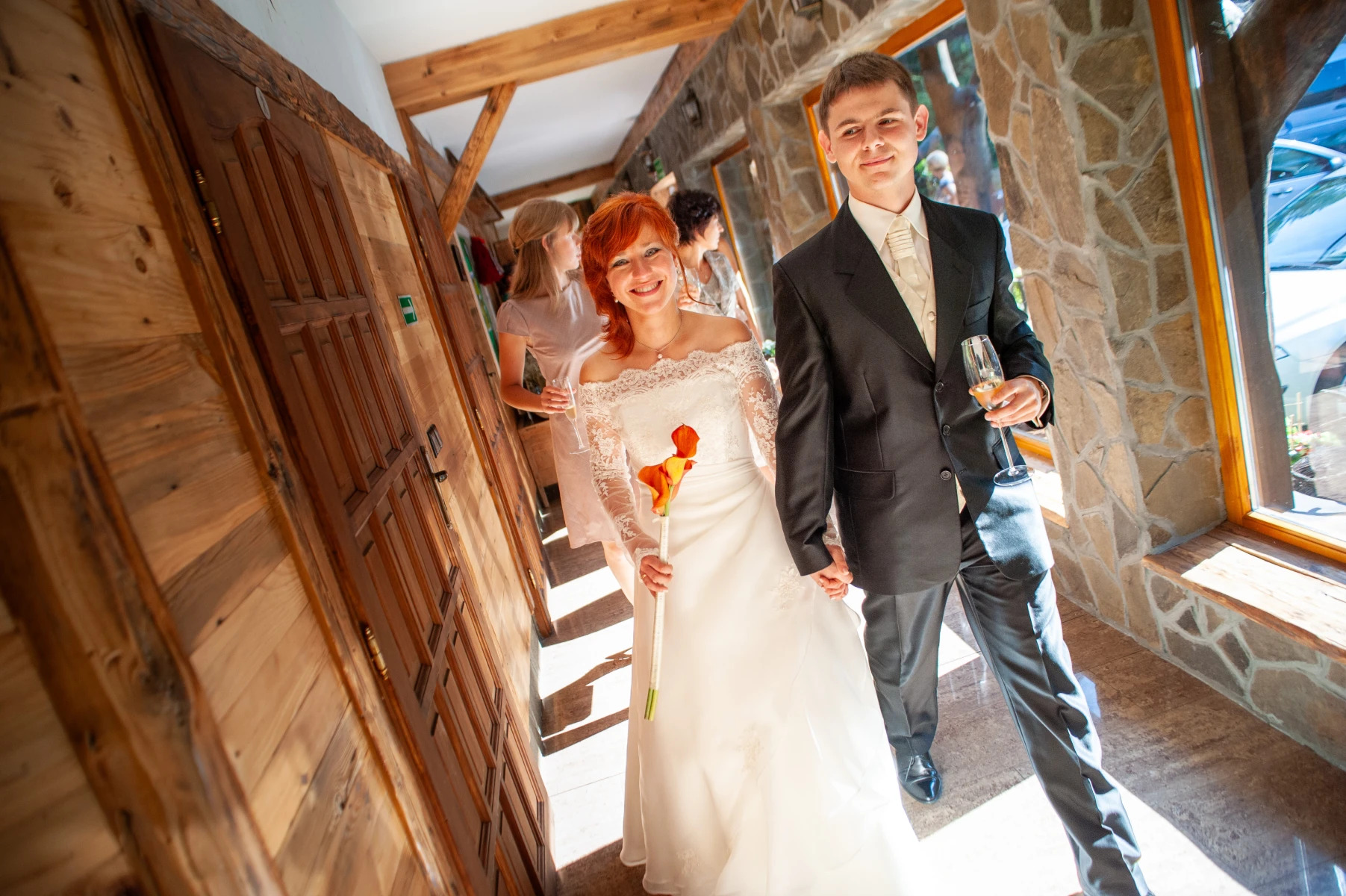 Para młoda w trakcie uroczystości, panna młoda w białej sukni z pomarańczową kalą, pan młody w czarnym garniturze trzyma kieliszek szampana, w tle goście i drewniane elementy wystroju.