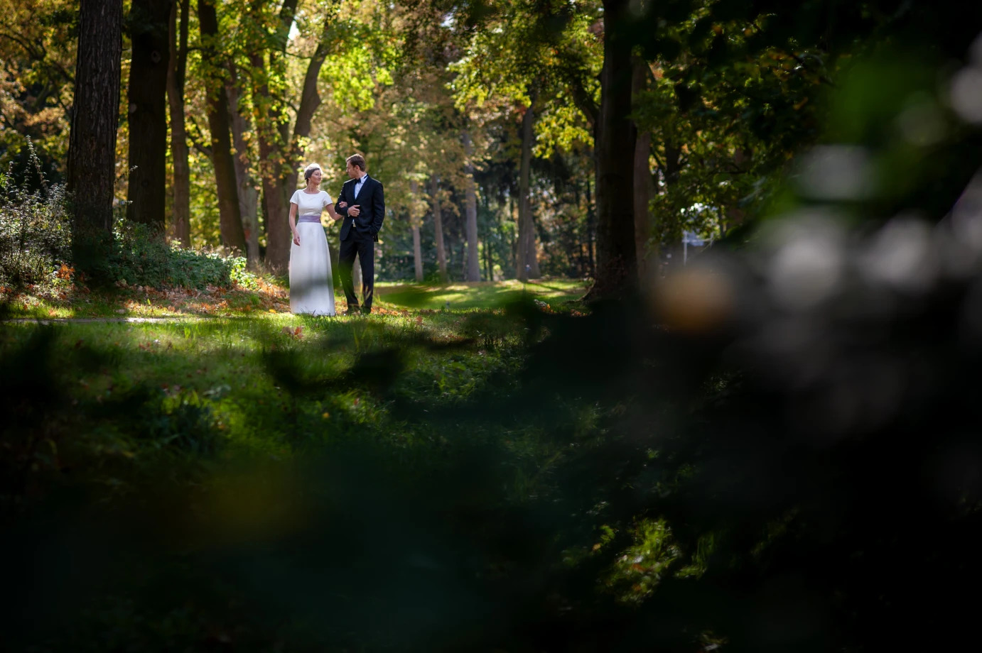 Para młoda spaceruje pośród drzew w słoneczny dzień, panna młoda w prostej sukni z fioletową szarfą, pan młody w czarnym garniturze, rozproszone światło słoneczne tworzy malowniczy efekt.