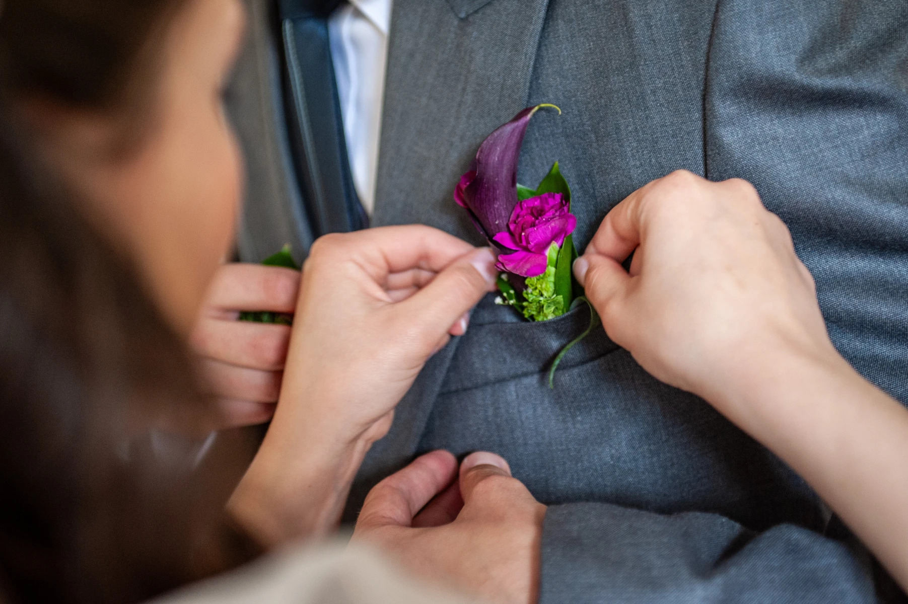 Osoba poprawia butonierkę z ciemnofioletowej kalii i różowej róży w klapie szarego garnituru.