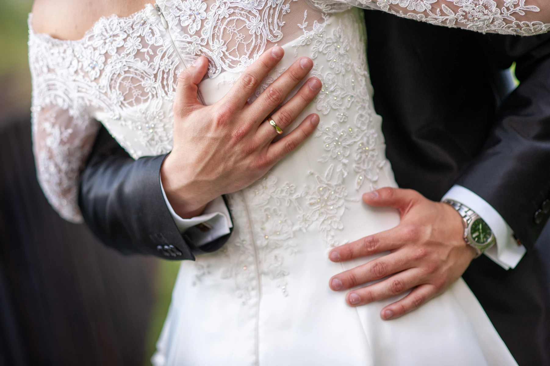 Pan młody obejmuje pannę młodą w koronkowej sukni, widoczny złoty pierścionek i zegarek na ręce.