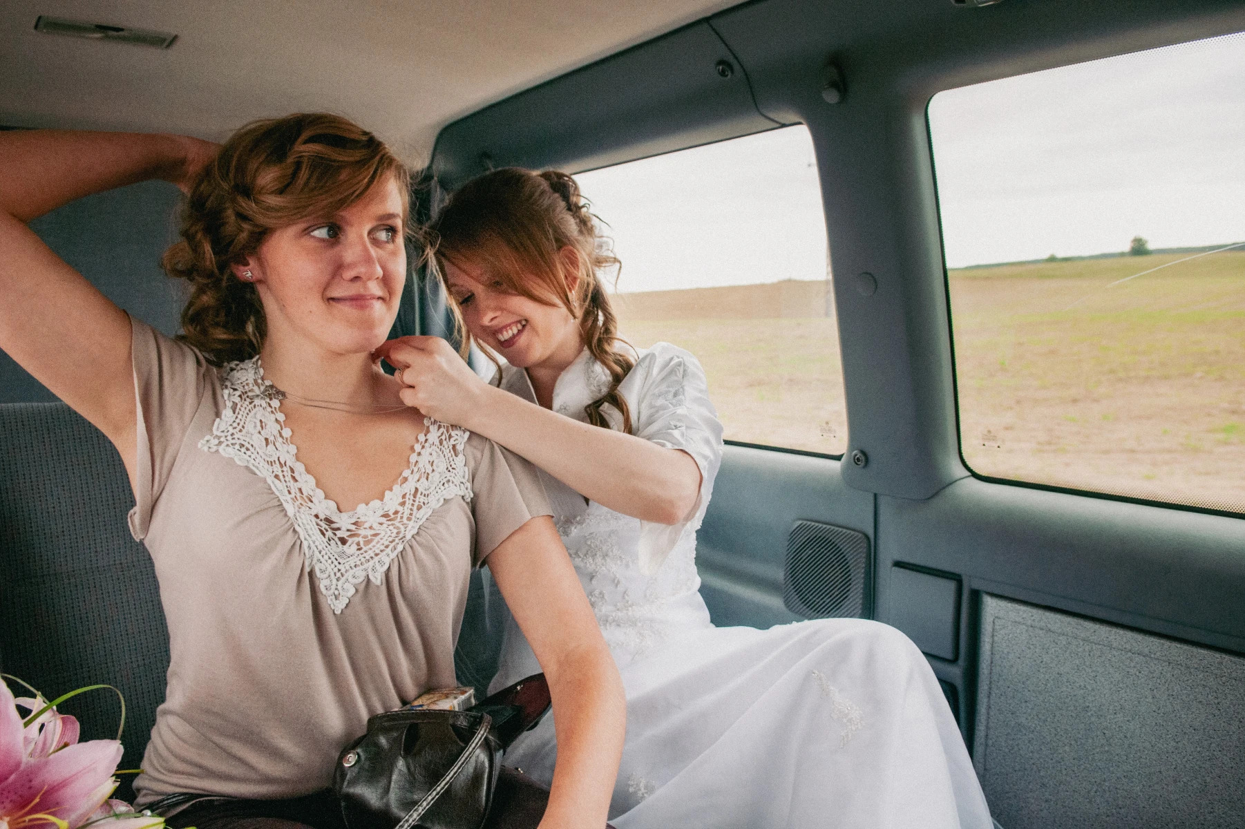 Panna młoda w sukni ślubnej, siedząca w samochodzie, zapina naszyjnik druhnie przed ceremonią, widok przez okno na pole.