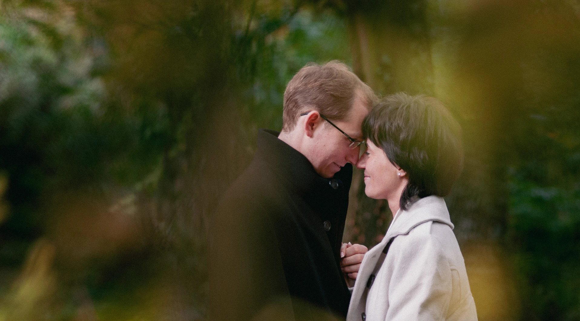 Para zakochanych, mężczyzna w czarnym płaszczu i okularach oraz kobieta w jasnym płaszczu, stykają się czołami na tle zieleni.