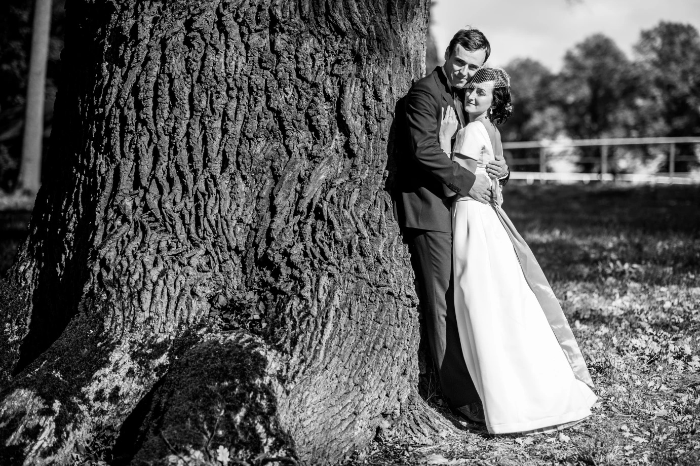 Czarno-białe zdjęcie pary młodej obejmującej się przy pniu starego, rozłożystego drzewa w parku, panna młoda w sukni z delikatną siateczką na włosach, pan młody w ciemnym garniturze.
