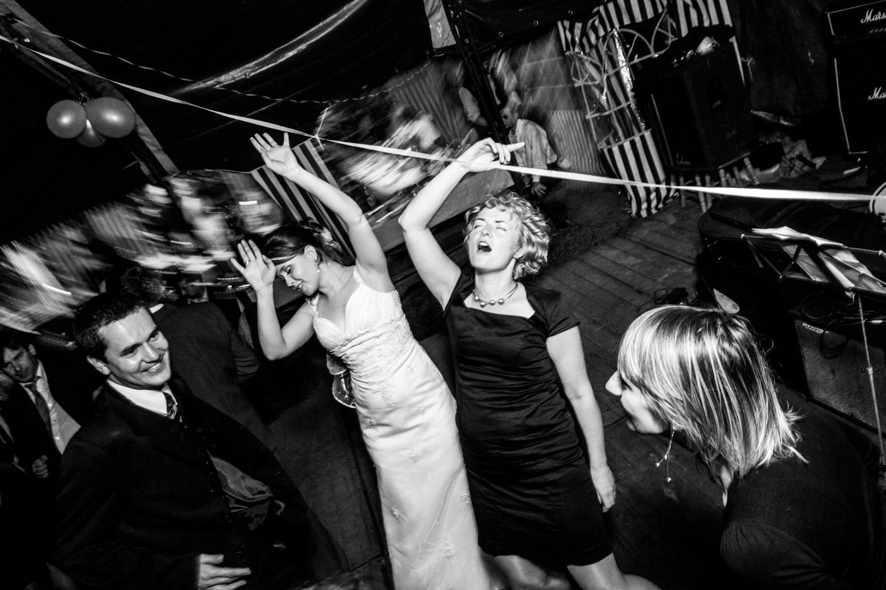 Czarno-białe zdjęcie z wesela: panna młoda w białej sukni i goście tańczą z podniesionymi rękami, w tle balony i dekoracje.
