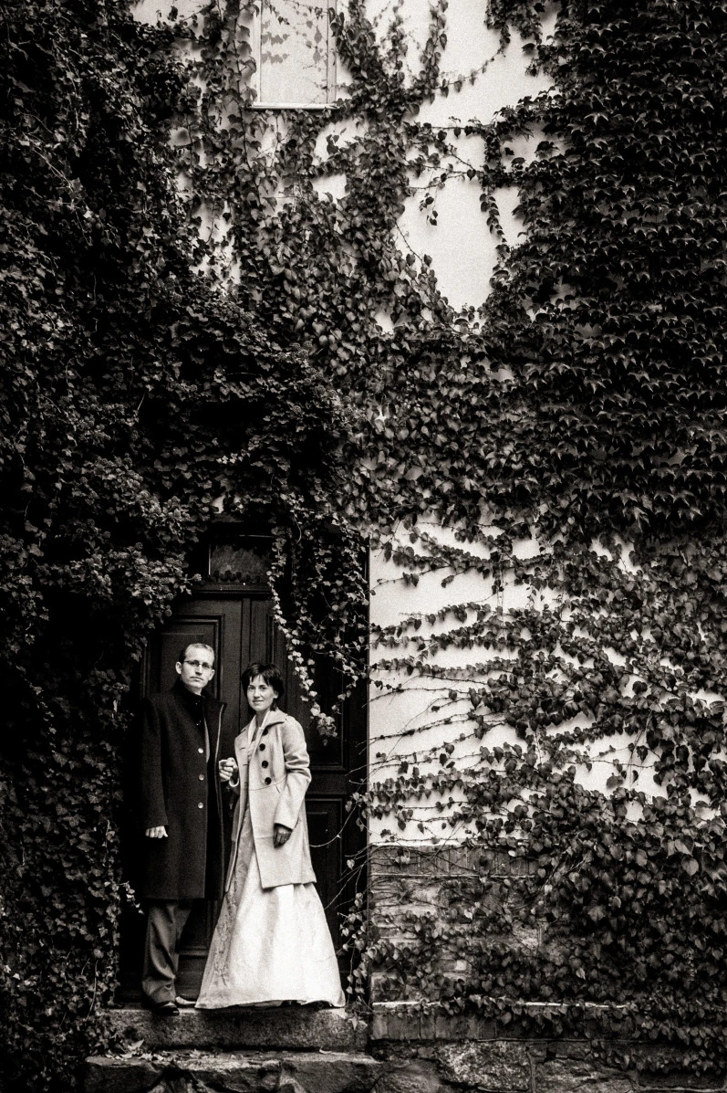 Czarno-białe zdjęcie pary narzeczeńskiej stojącej na schodach przed drzwiami obrośniętymi bluszczem, kobieta w sukni ślubnej i płaszczu, mężczyzna w ciemnym płaszczu.