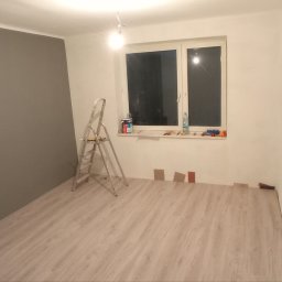 Mateusz Otwinowski Build & Design - Pokój w Lublinie w trakcie odświeżania: częściowo pomalowana szara ściana, drabina malarska, próbki paneli podłogowych i akcesoria malarskie na parapecie okna.