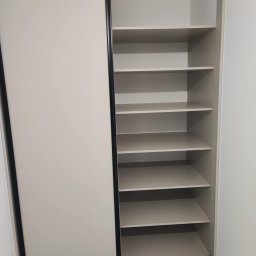 Mateusz Otwinowski Build & Design - Biała szafa wnękowa z przesuwnymi drzwiami i czarnymi obramowaniami, widok na puste półki. Minimalistyczny design, jasne wnętrze, marmurowa podłoga.