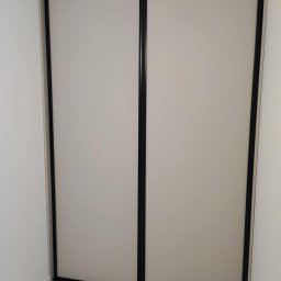 Mateusz Otwinowski Build & Design - Zamknięta szafa wnękowa z przesuwnymi drzwiami w kolorze jasnego beżu z czarnymi obramowaniami, widoczna na tle białej ściany i marmurowej podłogi.