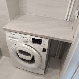 Mateusz Otwinowski Build & Design - Biała pralka Samsung z panelem sterowania pod jasnoszarym blatem, nad którym wisi biały kocioł gazowy z miedzianymi rurami. Całość w nowoczesnym wnętrzu z szarymi płytkami.