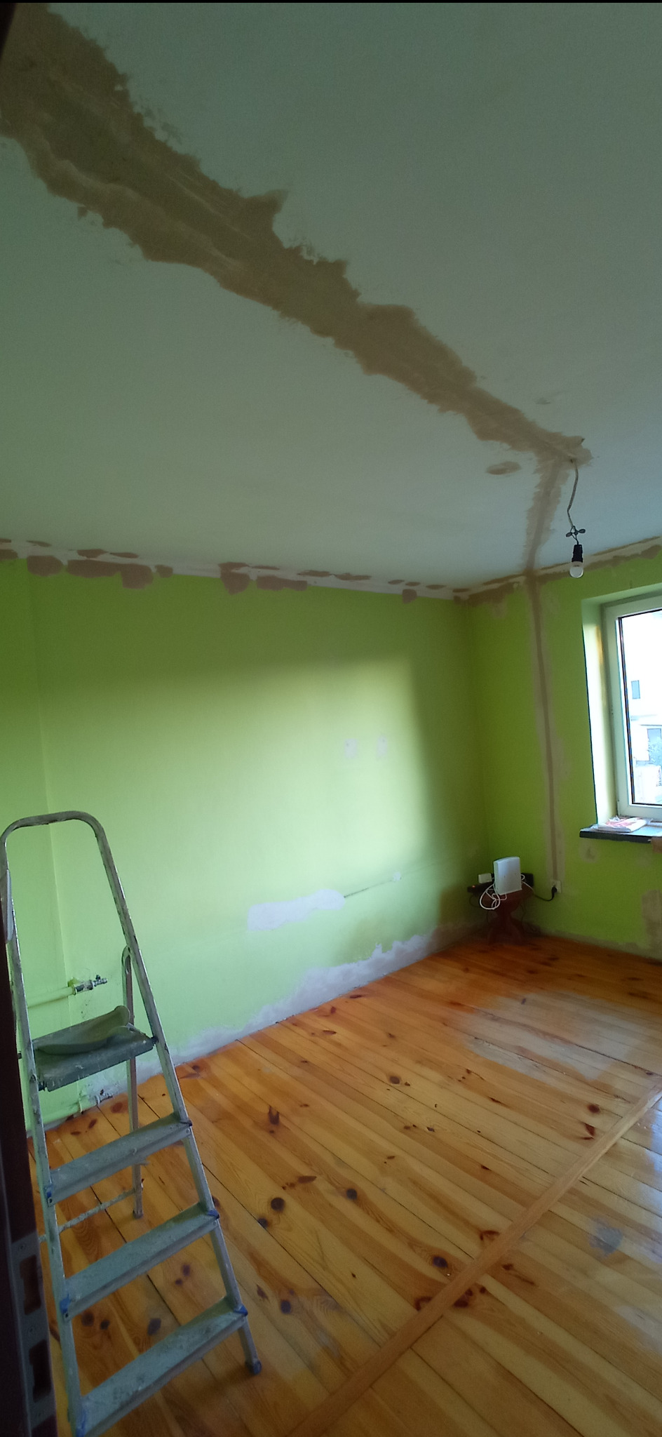 Pokój w trakcie prac przygotowawczych: zielone ściany z miejscowymi łatami, drewniana podłoga po cyklinowaniu, drabina aluminiowa, sufit z widocznymi śladami po szpachlowaniu, wisząca żarówka...