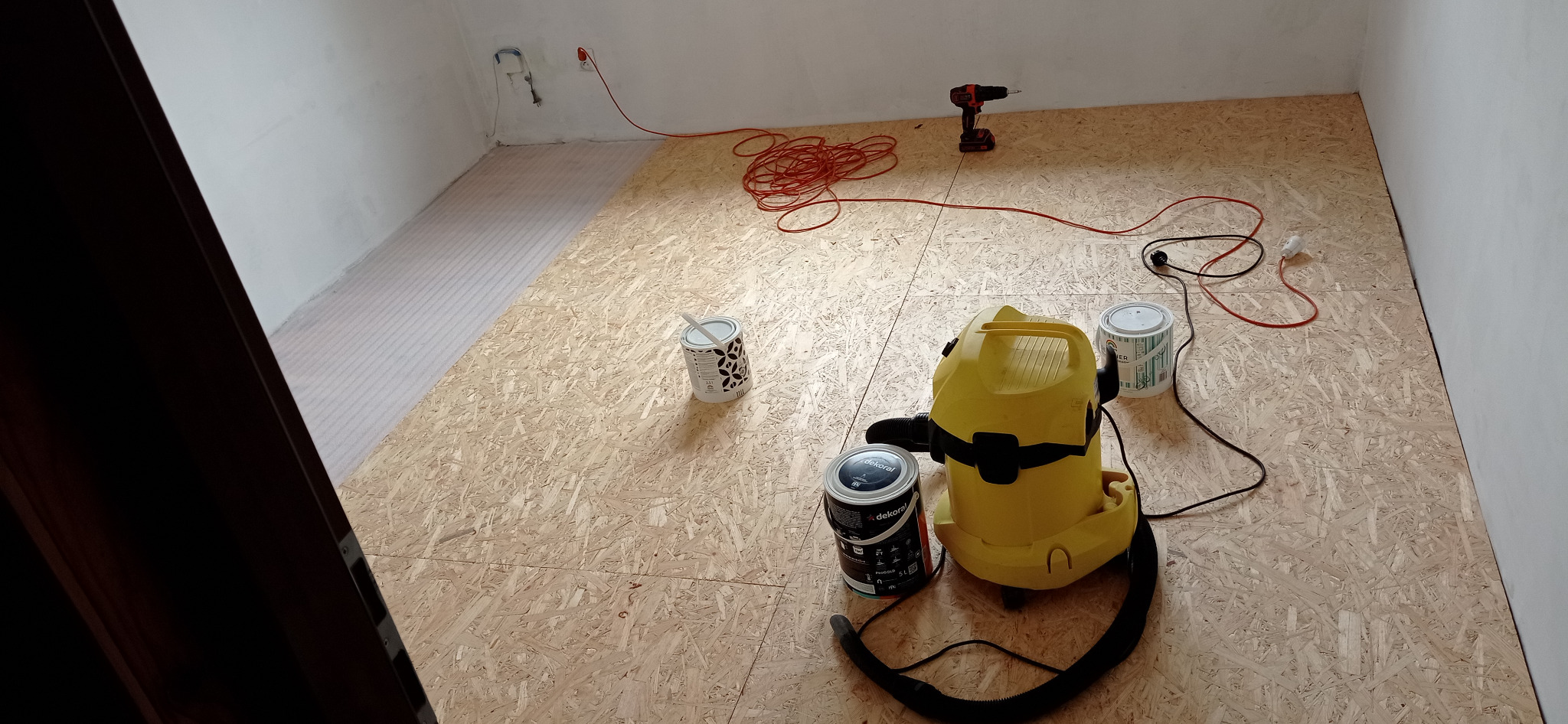 Przygotowanie do malowania podłogi: płyty OSB, puszki z farbą Dekoral, żółty odkurzacz przemysłowy i wkrętarka akumulatorowa na podłożu z folii izolacyjnej.