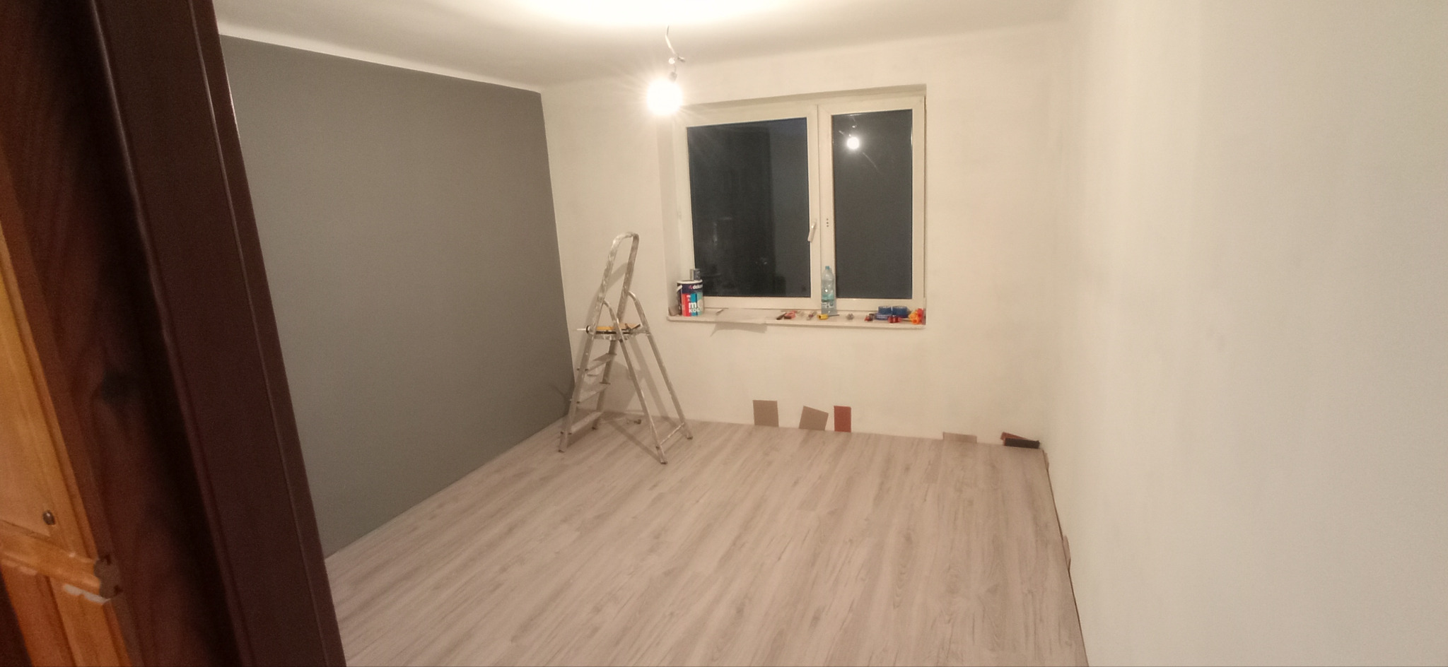 Pokój w Lublinie w trakcie odświeżania: częściowo pomalowana szara ściana, drabina malarska, próbki paneli podłogowych i akcesoria malarskie na parapecie okna.