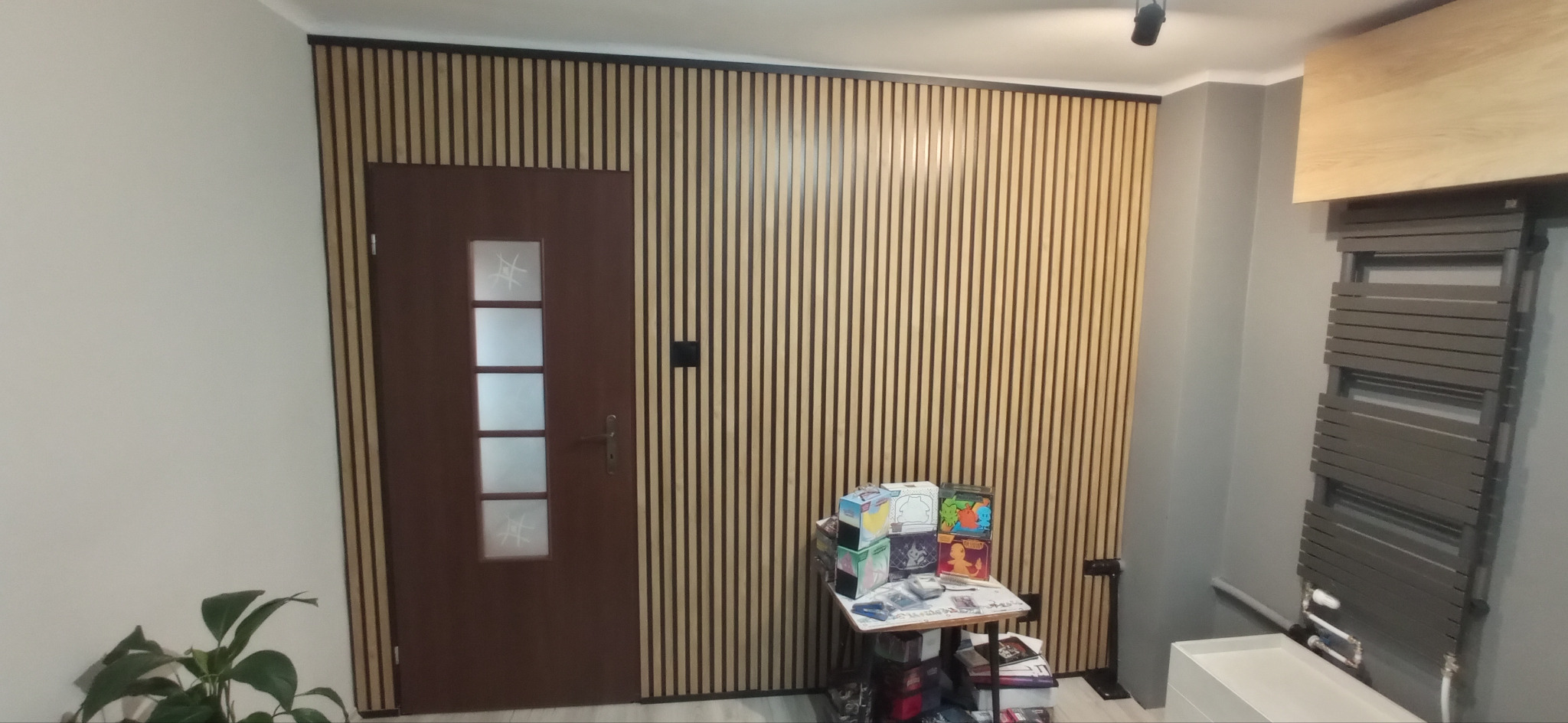 Panel ścienny z drewnianych listew w przedpokoju, obok brązowe drzwi z szybami i grzejnik drabinkowy, na dole widoczne rośliny i stoliczek z różnymi przedmiotami.