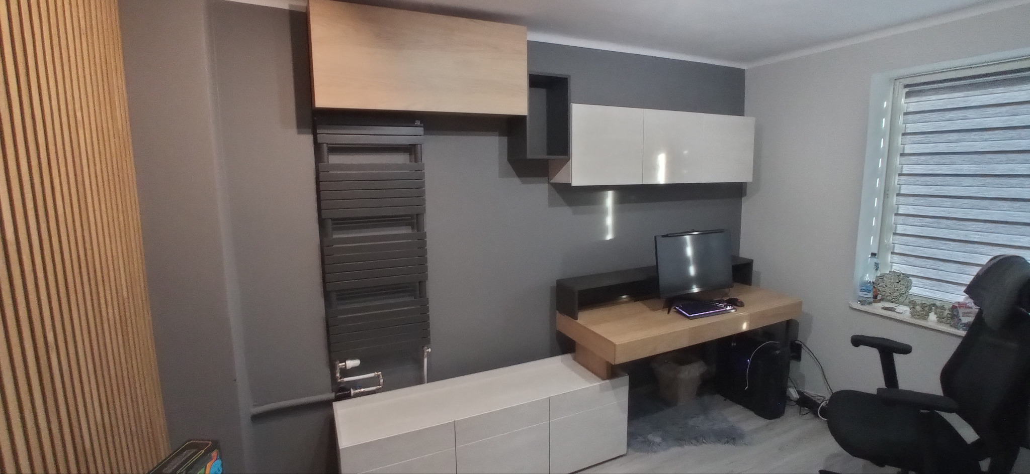 Szary grzejnik drabinkowy na ścianie obok drewnianych paneli ściennych i biurka z komputerem, w tle okno z żaluzjami.